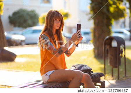 Videocall. Young smiling Caucasian tanned woman sitting on bench and using smartphone. Concept of psychology and online communication Videocall. Young smiling Caucasian tanned woman sitting on bench and using smartphone. Concept of psychology and online communication 127706887