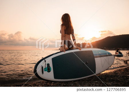 Sup boarding. A tanned young woman with a tattoo poses standing with a sup board. Back view. The concept of active lifestyle and surfing Sup boarding. A tanned young woman with a tattoo poses standing with a sup board. Back view. The concept of active lifestyle and surfing 127706895