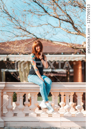 A young woman with tattoed hand sits on the balustrade and smiling. Vertical orientation. Outdoor 127706913