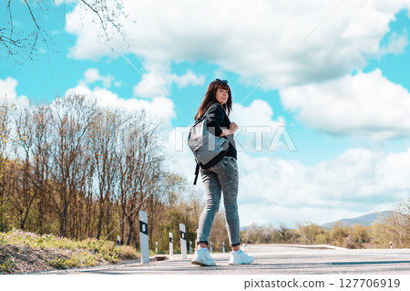A young pretty woman walks along the countryside road with a backpack in her hands. Back view. Copy space. Concept of hitchhiking and local travel 127706919