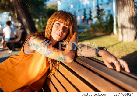 Portrait of young pretty smiling Caucasian woman with tattoos poses relax sitting on bench in sunny beach. Concept of psychology and mental health 127706924