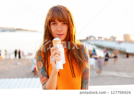Portrait of young smiling beautiful Caucasian woman with tattoos holds ice cream. Beach in background. Concept of freedom and summer vacation Portrait of young smiling beautiful Caucasian woman with tattoos holds ice cream. Beach in background. Concept of freedom and summer vacation 127706926