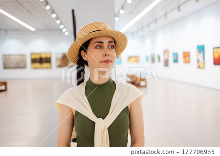 Portrait of a young elegantly dressed pretty woman student in a straw hat admiring the exhibition of paintings in the gallery. Contemporary art at the Art Museum 127706935