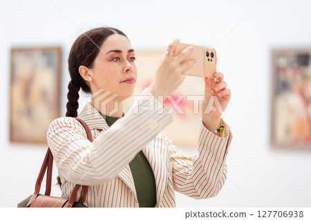 Portrait of young Caucasian beautiful woman in an elegant clothes takes photo with her smartphone in art gallery. Modern cultural education and online excursion Portrait of young Caucasian beautiful woman in an elegant clothes takes photo with her smartphone in art gallery. Modern cultural education and online excursion 127706938