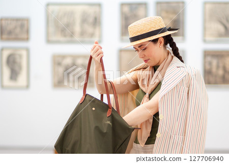 Mid shot of young adult Caucasian woman in a straw hat and elegant clothes visit modern art gallery. Woman uses her bag. Paintings in background. Concept of Museum Day and cultural education Mid shot of young adult Caucasian woman in a straw hat and elegant clothes visit modern art gallery. Woman uses her bag. Paintings in background. Concept of Museum Day and cultural education 127706940