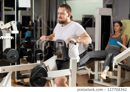 Bearded man in gym performs exercises for arm muscles using simulators and fitness center equipment. 127708362