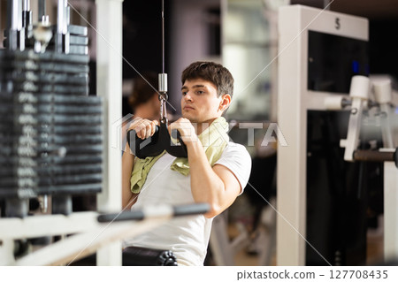 Guy in gym performs exercises for arm muscles using cable machine and pull weighted rope Guy in gym performs exercises for arm muscles using cable machine and pull weighted rope 127708435