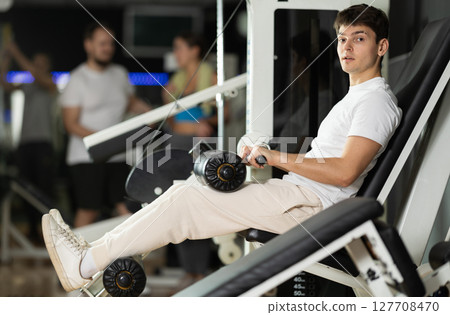 Young man in gym performs exercises for leg muscles using simulators and fitness center equipment. 127708470