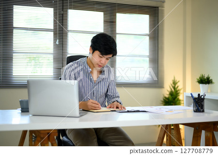 Man focused on planning or journaling beside a laptop in a bright, modern workspace 127708779