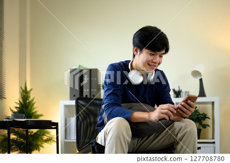 Smiling young man using smartphone with headphones at home 127708780