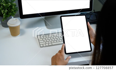 Close up of hands holding blank screen tablet in a modern workspace with coffee, and computer monitor 127708857