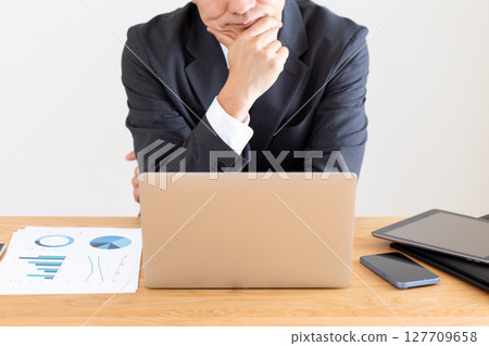 A middle-aged male businessman in a suit worried while working on a computer at his desk 127709658