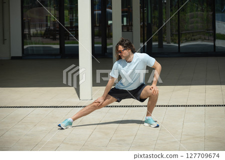 Fit brunette man wearing sports casual clothes doing stretching and warm up exercise on urban outdoor public park. Summer activity. Healthy lifestyle. 127709674