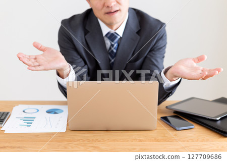 A middle-aged male businessman in a suit posing with his hands up while working on a computer at his desk A middle-aged male businessman in a suit posing with his hands up while working on a computer at his desk 127709686
