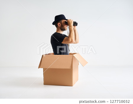 Man in hat looking through binoculars sitting inside cardboard box. Man in hat looking through binoculars sitting inside cardboard box. 127710032