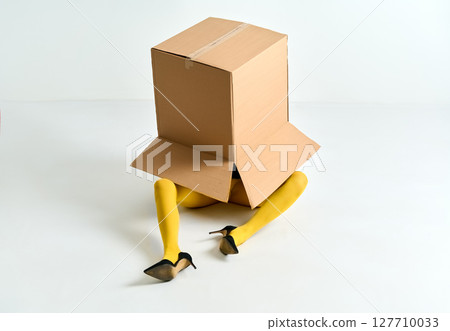 Woman in yellow tights and black high heels hidden under oversized cardboard box on white floor Woman in yellow tights and black high heels hidden under oversized cardboard box on white floor 127710033