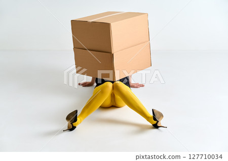 Woman in yellow tights and black high heels hidden under oversized cardboard box on white floor Woman in yellow tights and black high heels hidden under oversized cardboard box on white floor 127710034