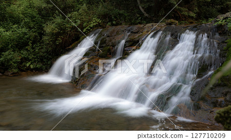 This tranquil waterfall flows gracefully over rocks, with vibrant green plants framing the scene. 127710209