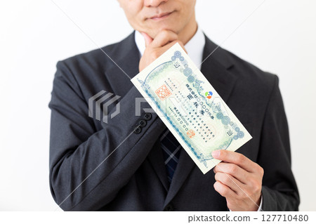 A middle-aged male businessman in a suit worried about holding a stock certificate 127710480