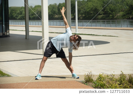 Fit brunette man wearing sports casual clothes doing stretching and warm up for arms on urban outdoor public park. Summer activity. Healthy lifestyle. 127711033