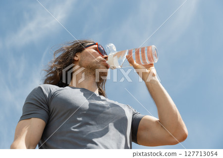 Handsome curly brunette man wear gray t-shirt drinking water bottle against blue sky background. Summer heat weather. Outdoors. Hero view. Close up. 127713054