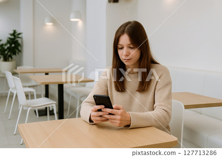 Young caucasian woman sitting in a modern white cafe using smartphone for online communication. concept of digital connectivity, leisure time, minimalist decor Young caucasian woman sitting in a modern white cafe using smartphone for online communication. concept of digital connectivity, leisure time, minimalist decor 127713286