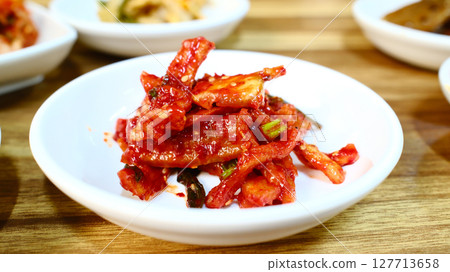Spicy Korean dried squid banchan with red chili seasoning served as traditional seafood side dish with intense umami flavor Spicy Korean dried squid banchan with red chili seasoning served as traditional seafood side dish with intense umami flavor 127713658