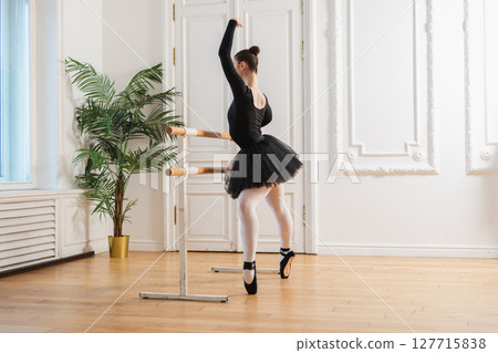 Young ballerina in tutu skirt stretching practicing ballet positions dancing near ballet barre in dance class. Young classical dancer woman in dance studio. Ballet class training Ballet school concept 127715838