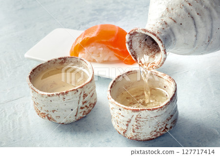 Sake poured into ochoko, traditional cup, with salmon nigiri in the background Sake poured into ochoko, traditional cup, with salmon nigiri in the background 127717414