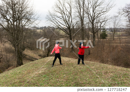 Two individuals engaged in a playful dance on a grassy hilltop at dusk, surrounded by bare trees and the serene beauty of autumn's end in a rural landscape 127718247