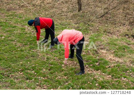 Two individuals actively foraging for wild plants in a lush green field surrounded by trees on a sunny day, exploring nature's hidden treasures with curiosity 127718248
