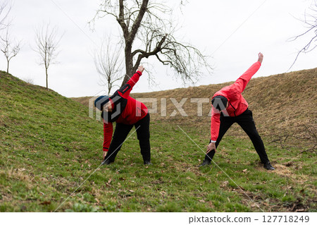 Stretching outdoors in early spring amidst bare trees and a gentle hillside, two individuals engage in a morning warm up routine, embracing wellness and nature 127718249