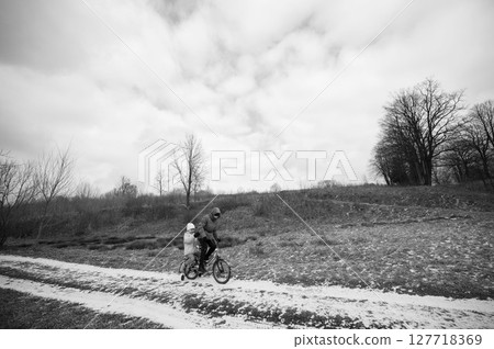 Cycling adventure through a serene landscape with a parent and child enjoying a quiet ride on a winding path surrounded by barren trees on a cloudy day 127718369