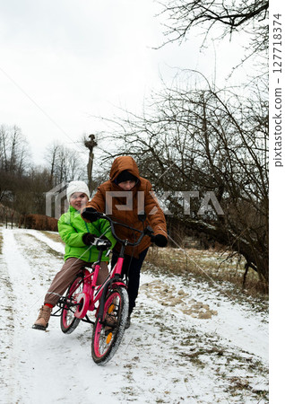 Two joyful children riding a bright pink bicycle together on a snowy path in winter, surrounded by barren trees and a tranquil landscape 127718374