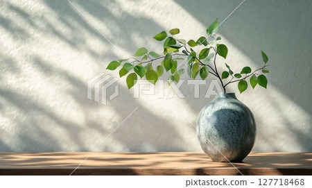 A simple green branch placed in a textured stone vase on a wooden table, illuminated by soft natural light, representing serenity and minimalist beauty A simple green branch placed in a textured stone vase on a wooden table, illuminated by soft natural light, representing serenity and minimalist beauty 127718468