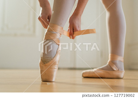 Ballerina puts on pointe shoes on leg in dance class. Classical ballet dancer woman in dance studio. Ballerina putting on tying classical pointe shoes for dance training. Ballet school concept 127719002