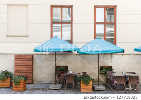 Charming Outdoor Cafe Scene with Blue Canopies, Elegant Tables, and Stylish Planters Set Against a Neutral Wall Offering a Cozy Dining Experience in a European City Charming Outdoor Cafe Scene with Blue Canopies, Elegant Tables, and Stylish Planters Set Against a Neutral Wall Offering a Cozy Dining Experience in a European City 127719323