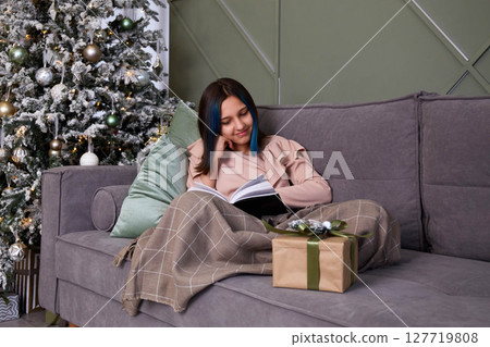 A cute girl is reading a book on the sofa, covered with a blanket. Next to the Christmas tree with toys and gifts. 127719808