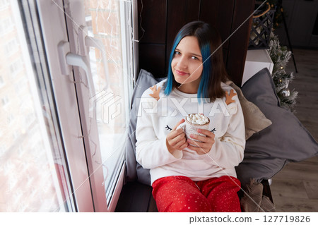 A pretty girl in pajamas is sitting on the windowsill with a cup of cocoa with marshmallows. 127719826