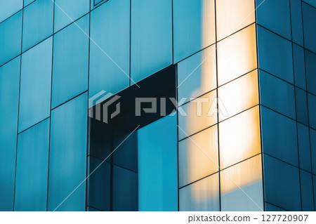 Modern building facade with reflective glass panels and geometric design, featuring sunlight creating warm glow on surface 127720907