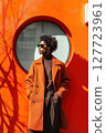 Stylish man standing against vibrant orange wall with circular window during sunny day in urban environment 127723961