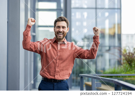 Mature man celebrates success outside modern building. Wearing casual shirt, he expresses joy with raised arms. Bright smile reflects happiness and achievement. Urban setting enhances mood. Mature man celebrates success outside modern building. Wearing casual shirt, he expresses joy with raised arms. Bright smile reflects happiness and achievement. Urban setting enhances mood. 127724194