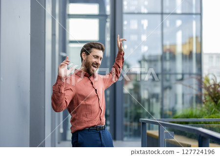 Confident man expressing joy outdoors near office after achieving goal or success. Wearing casual shirt with a vibrant smile, showcasing business or career milestone enthusiasm. 127724196