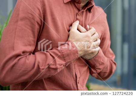 Person holding chest in rust-colored shirt signifies pain, stress, or heart-related concern. Close-up emphasizes emotion and tension in hands. Suitable for health or wellness themes. Person holding chest in rust-colored shirt signifies pain, stress, or heart-related concern. Close-up emphasizes emotion and tension in hands. Suitable for health or wellness themes. 127724220