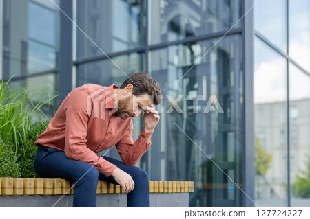 Mature businessman in city street sitting outside office building, showing stress and contemplation. Professional adult expressing concern about work, dressed casual, highlighting urban lifestyle. Mature businessman in city street sitting outside office building, showing stress and contemplation. Professional adult expressing concern about work, dressed casual, highlighting urban lifestyle. 127724227