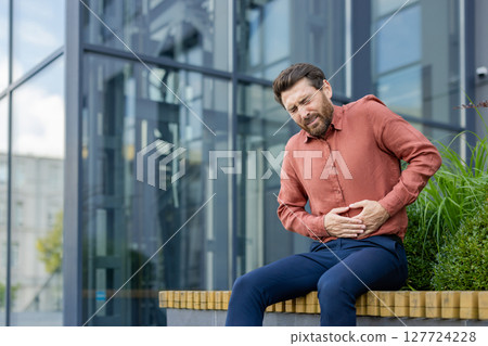 A man in a red shirt is sitting on a bench with a stomach ache. He is holding his stomach and he is in pain A man in a red shirt is sitting on a bench with a stomach ache. He is holding his stomach and he is in pain 127724228