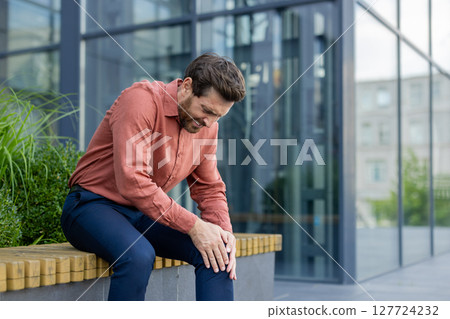 Mature male professional sitting outdoors in city showing signs of knee pain. Man wearing casual shirt and trousers holds knee, indicating discomfort or injury. 127724232