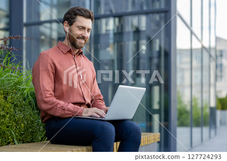 Business professional using laptop outdoors, wearing headphones, enjoying modern technology. Smiling and relaxed, he sits in natural setting by glass building, showcasing productivity. 127724293