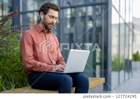 Mature businessman sits outside using laptop, with headphones, displaying remote work lifestyle. Casual attire suggests comfort and focus, embodying work life balance through technology interaction. 127724295