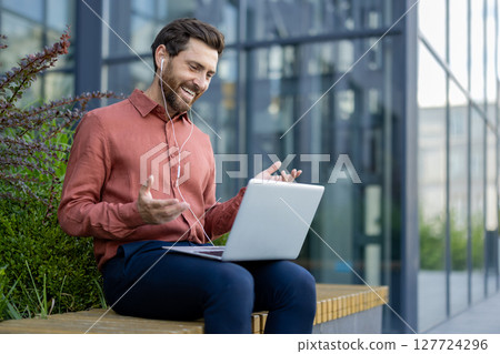 Businessman using laptop for digital meeting outdoors. Smiling in casual attire with earphones, seated on bench with modern building background. Depicts communication, technology, success. 127724296